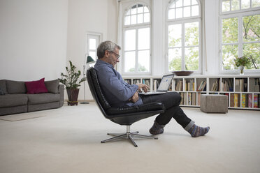 Älterer Mann sitzt zu Hause auf einem Stuhl und benutzt einen Laptop - RBF05373