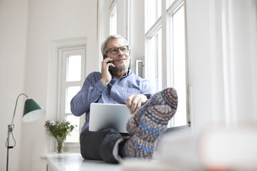Mature man at home sitting at the window using tablet and cell phone - RBF05381