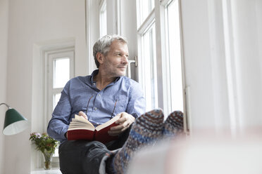 Entspannter reifer Mann zu Hause mit Buch und Blick aus dem Fenster - RBF05382
