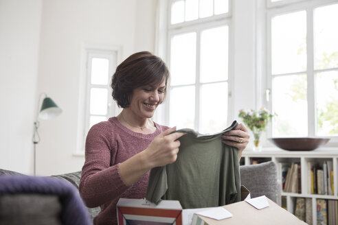 Smiling woman at home sitting on the sofa looking at garment - RBF05395