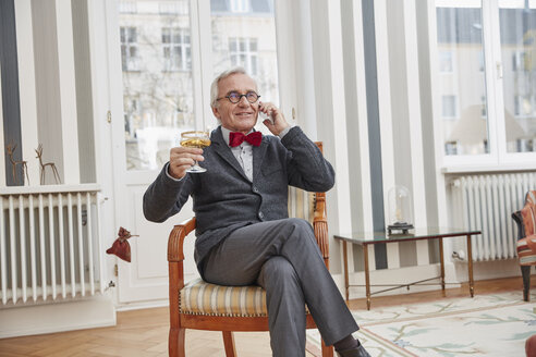 Lächelnder älterer Mann am Telefon auf einem Stuhl sitzend mit einem Sektglas - RHF01723