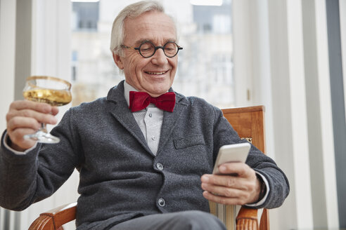 Lächelnder älterer Mann mit Handy sitzt auf einem Stuhl und hält ein Sektglas - RHF01725