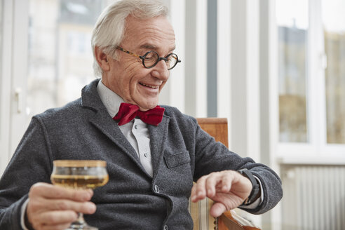 Lächelnder älterer Mann, der auf einem Stuhl sitzt und ein Sektglas hält und auf seine Smartwatch schaut - RHF01728