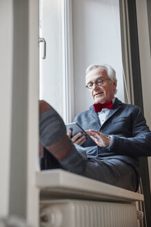 Älterer Mann sitzt auf der Fensterbank und benutzt ein Tablet - RHF01737