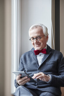 Lächelnder älterer Mann sitzt auf der Fensterbank und benutzt ein Tablet - RHF01739