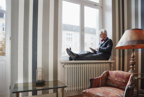 Älterer Mann sitzt auf der Fensterbank und benutzt ein Tablet - RHF01742