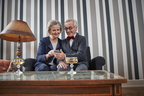 Glückliches älteres Paar sitzt mit Smartphone auf der Couch - RHF01773