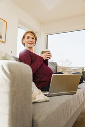 Pregnant woman with laptop on couch holding mug - UUF09586