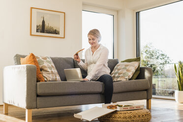 Smiling pregnant woman on couch holding a slice of pizza using laptop - UUF09591