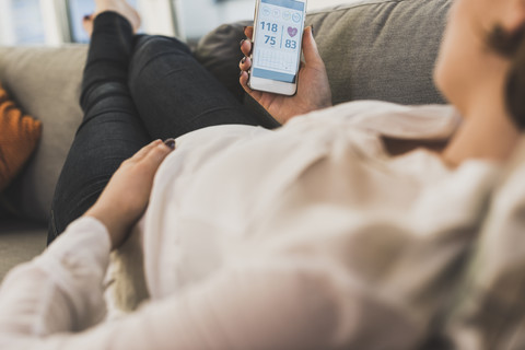 Schwangere Frau auf der Couch, die mit ihrem Smartphone Gesundheitsdaten überprüft, lizenzfreies Stockfoto