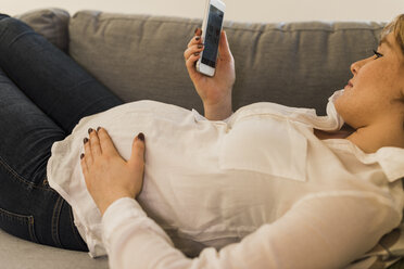 Schwangere Frau auf der Couch, die mit ihrem Smartphone Gesundheitsdaten überprüft - UUF09597