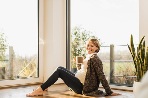 Smiling pregnant woman sitting on floor enjoying a drink in mug stock photo