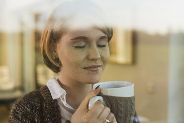 Young woman behind windowpane holding a drink in mug - UUF09612