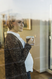 Schwangere Frau hinter einer Fensterscheibe, die ein Getränk in einem Becher hält - UUF09613