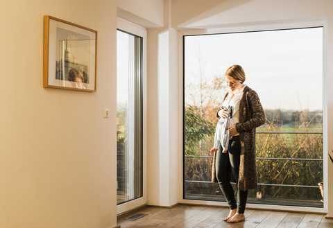 Schwangere Frau steht am Fenster und hält Babykleidung, lizenzfreies Stockfoto