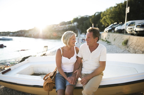 Älteres Paar sitzt am Rande eines Bootes am Strand in der Abenddämmerung, lizenzfreies Stockfoto