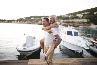 Senior man giving his wife a piggyback ride on a jetty - HAPF01279