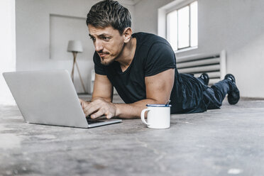 Mann auf dem Boden liegend mit Laptop in leerem Loft - KNSF00839