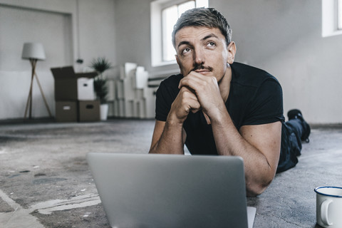 Mann auf dem Boden liegend mit Laptop in leerem Dachboden, lizenzfreies Stockfoto