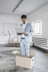 Geschäftsmann mit Laptop in einem Karton auf einem leeren Dachboden - KNSF00850