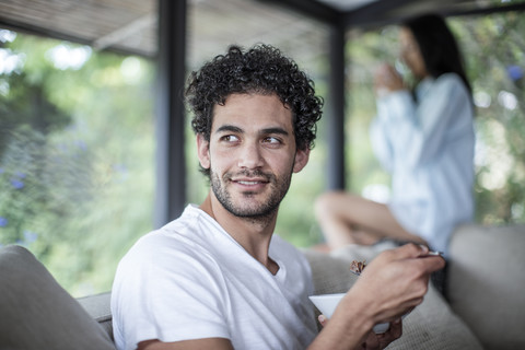 Junger Mann isst Müsli zu Hause mit Frau im Hintergrund, lizenzfreies Stockfoto