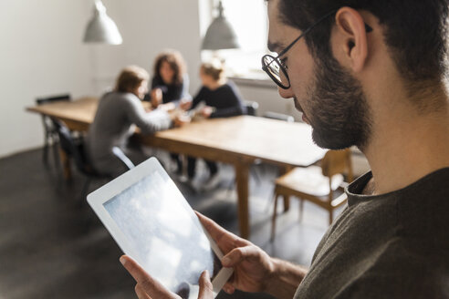 Junger Mann, der ein digitales Tablet in einem modernen Büro benutzt, mit Kollegen am Tisch im Hintergrund - TCF05283