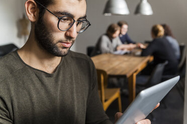 Junger Mann, der ein digitales Tablet in einem modernen Büro benutzt, mit Kollegen am Tisch im Hintergrund - TCF05284