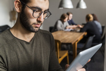 Young man using digital tablet in modern office with coworkers at table in background - TCF05284