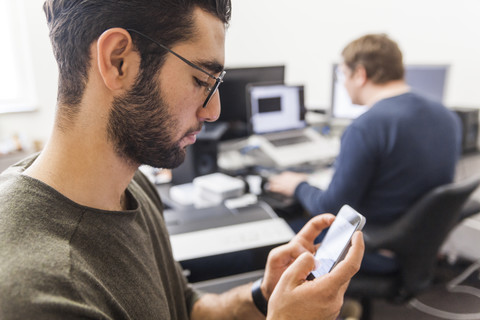 Junger Mann benutzt Smartphone in modernem Büro mit Mitarbeiter im Hintergrund, lizenzfreies Stockfoto