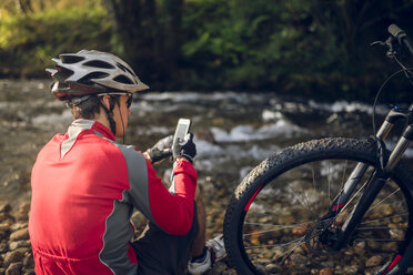 Mountainbiker sitzt am Fluss und benutzt sein Smartphone - RAEF01623