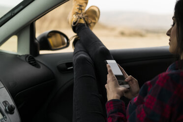 Beine einer jungen Frau, die sich aus dem Autofenster lehnt, während sie ihr Mobiltelefon benutzt - KKAF00266