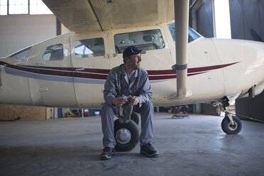 Mechanic sitting next to light aircraft in airfield hangar - ZEF12235