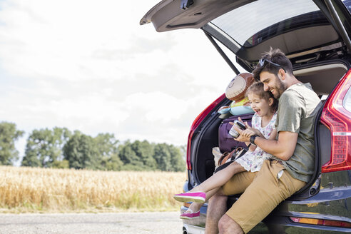 Father and daughter sitting in open car boot looking at camera at break of a road trip - WESTF22377