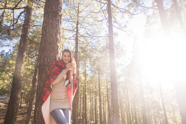 Lächelnde junge Frau mit Decke und Getränk an einen Baumstamm gelehnt im Wald - SIPF01231