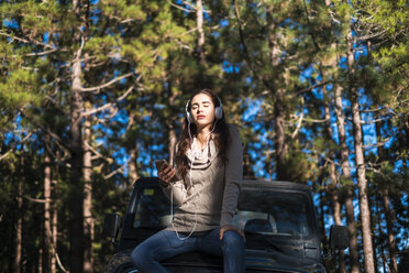 Junge Frau mit geschlossenen Augen sitzt auf der Motorhaube eines Autos im Wald und hört Musik mit Kopfhörern - SIPF01236