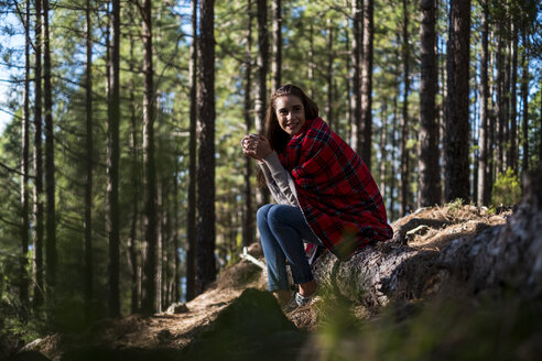 Smiling young woman with blanket and beverage sitting on rock in the forest - SIPF01237