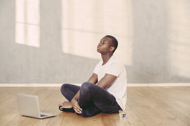 Daydreaming young man sitting on floor with laptop and glass of water - FMKF03386