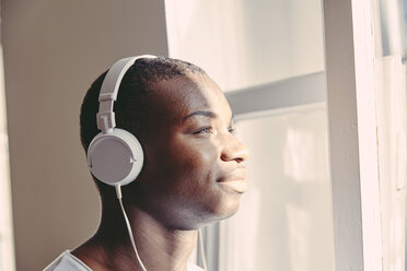 Portrait of smiling young man listening music with headphones while looking through window - FMKF03387