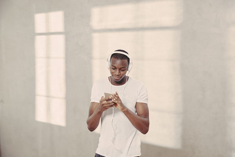 Lächelnder junger Mann, der mit Kopfhörern Musik hört und auf sein Mobiltelefon schaut, lizenzfreies Stockfoto