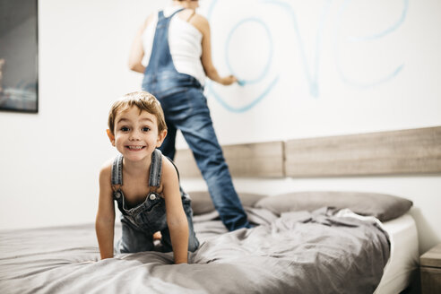 Porträt eines kleinen Jungen, der auf dem Bett spielt, während seine Mutter die Wand bemalt - JRFF01126