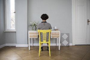 Rückenansicht einer jungen Frau, die auf einem gelben Stuhl am Schreibtisch zu Hause sitzt - RBF05432