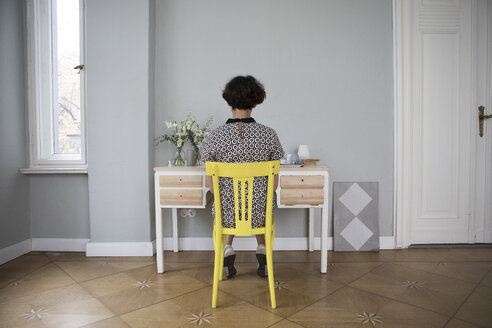 Back view of young woman sitting on yellow chair at desk at home - RBF05432