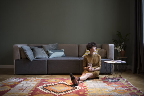 Young woman sitting on the floor in her living room looking through window - RBF05443