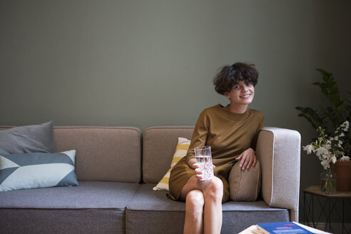 Porträt einer lächelnden jungen Frau mit einem Glas Wasser, die zu Hause auf der Couch sitzt - RBF05444