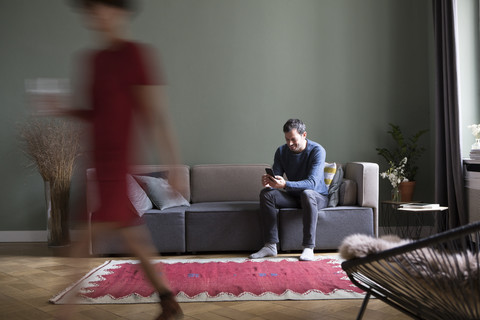 Mann sitzt auf der Couch im Wohnzimmer, während seine Freundin im Vordergrund vorbeigeht, lizenzfreies Stockfoto