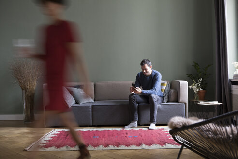 Mann sitzt auf der Couch im Wohnzimmer, während seine Freundin im Vordergrund vorbeigeht - RBF05453