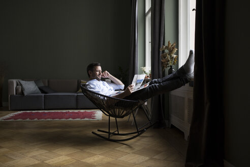 Mann entspannt sich auf einem Schaukelstuhl in seinem Wohnzimmer und liest Zeitung - RBF05479