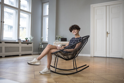 Porträt einer lächelnden jungen Frau, die zu Hause auf einem Schaukelstuhl sitzt - RBF05501
