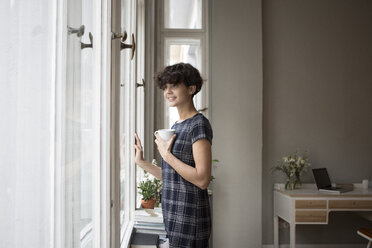 Porträt einer lächelnden jungen Frau, die durch das Fenster ihres Hauses schaut - RBF05505