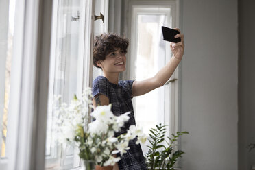 Lächelnde junge Frau steht vor einem Fenster und macht ein Selfie mit ihrem Smartphone - RBF05508